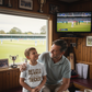 Man and child watching cricket on TV in a room with sports memorabilia.  The young boy is wearing a Cows Corner t-shirt with the funny and sweet cricket slogan on the front that reads 'Dad’s Like a Spin Bowler, Always Full of Tricks!' 