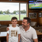 Man and young boy sitting together in a sports bar with a TV showing a cricket match.  The young boy is wearing a Cows Corner t-shirt with the funny and sweet saying on the front that reads 'Dads Like A Good Yorker, Keeps Me On My Toes'