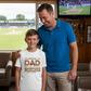 Man and young boy standing together in a room with a sports field view, the boy wearing a t-shirt with a humorous message.  The young boy is wearing a Cows Corner hoodie with the funny and sweet cricket slogan on the front that reads 'I'd Appeal to Dad, But He Never Changes His Decision'. Cows Corner gifts are perfect for sport-mad fans who love a bit of humour and personality in their presents. They’re ideal for friends, partners, parents, coaches, teammates, and proud mums and dads who live and breath
