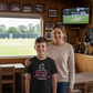 Woman and child standing together in a room with a sports field view, smiling at the camera.  The young boy is wearing a Cows Corner t-shirt with the funny and sweet cricket saying on the front that reads 'Mum's My Personal Third Umpire, Always Reviewing My Choices 