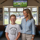 Woman and young boy standing together in a room with a TV showing a cricket match.  The young boy is wearing a Cows Corner t-shirt with the funny and sweet cricket saying on the front that reads 'Mums Like A Good Bat Grip, Always Holding Things Together'