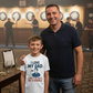 Man and child standing together in a pub setting with dartboards in the background. The boy is wearing a Cows Corner t-shirt with the funny darts slogan 'I Love My Dad… Even If He Can’t Hit A Double'.  Cows Corner gifts are perfect for sport-mad fans, these gifts work brilliantly for birthdays, new baby celebrations, Father’s Day, Mother’s Day, Christmas, anniversaries, thank you gifts, end-of-season team awards, graduations, retirements, and just-because moments when you want to raise a smile. 
