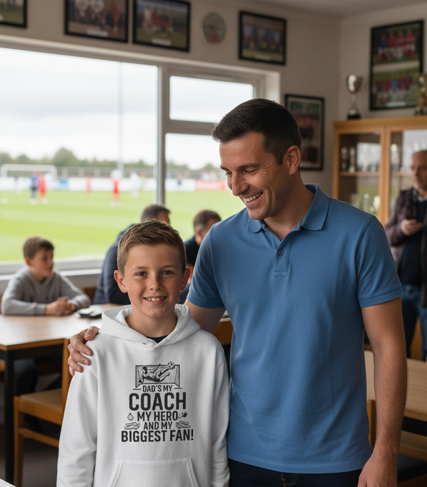 Man and young boy standing together in a sports setting with a classroom background. The boy is wearing a Cows Corner hoodie with the funny football slogan 'Dad’s My Coach My Hero and My Biggest Fan!'. Cows Corner gifts are perfect for sport-mad fans, these gifts work brilliantly for birthdays, new baby celebrations, Father’s Day, Mother’s Day, Christmas, anniversaries, thank you gifts, end-of-season team awards, graduations, retirements, and just-because moments when you want to raise a smile. 
