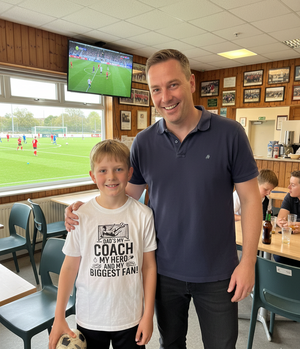 Man and young boy standing together in a sports facility with a TV showing a soccer match.  The boy is wearing a Cows Corner T-shirt with the funny and sweet football slogan on the front that reads 'Dad’s My Coach My Hero and My Biggest Fan!'. Cows Corner gifts are perfect for sport-mad fans, these gifts work brilliantly for birthdays, new baby celebrations, Father’s Day, Mother’s Day, Christmas, anniversaries, thank you gifts, end-of-season team awards, graduations, retirements, and just-because moments