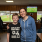 Woman and child standing together in a sports facility with a television screen showing a football match.  The boy is wearing a Cows Corner T-shirt with the funny and sweet football slogan on the front that reads 'Born To Play Football Raised by a Football Mum!'. Cows Corner gifts are perfect for sport-mad fans, these gifts work brilliantly for birthdays, new baby celebrations, Father’s Day, Mother’s Day, Christmas, anniversaries, thank you gifts, end-of-season team awards, graduations, retirements