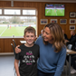 Woman and child in a sports facility with a TV screen showing a soccer match.  The boy is wearing a Cows Corner T-shirt with the funny and sweet slogan on the front that reads 'Mum Shouts at the TV Like She’s on the Team!'  Cows Corner gifts are perfect for sport-mad fans, these gifts work brilliantly for birthdays, new baby celebrations, Father’s Day, Mother’s Day, Christmas, anniversaries, thank you gifts, end-of-season team awards, graduations, retirements, and just-because moments 