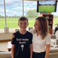 Woman and child in a living room with a TV showing golf on a screen. The boy is wearing a Cows Corner t-shirt that has a funny and sweet golf saying on the front that reads 'Best Mum by Par'. Cows Corner gifts are perfect for sport-mad fans, these gifts work brilliantly for birthdays, new baby celebrations, Father’s Day, Mother’s Day, Christmas, anniversaries, thank you gifts, end-of-season team awards, graduations, retirements, and just-because moments when you want to raise a smile. 
