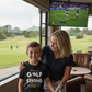 Woman and child in a golf club lounge watching a game on television. The boy is wearing a Cows Corner t-shirt with the funny and sweet golf saying on the front that reads 'Mum’s Golf Game Is As Strong as Her Coffee!'. Cows Corner gifts are perfect for sport-mad fans, these gifts work brilliantly for birthdays, new baby celebrations, Father’s Day, Mother’s Day, Christmas, anniversaries, thank you gifts, end-of-season team awards, graduations, retirements, and just-because moments when you want to raise a smi