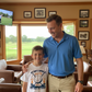Man and young boy standing together in a golf-themed room with a TV screen showing a golf course. The boy is wearing a Cows Corner t-shirt with a funny and sweet golf slogan on the front that reads 'My Dad’s Swing Is Smooth but His Dad Jokes Are Rough!'  Cows Corner gifts are perfect for sport-mad fans, these gifts work brilliantly for birthdays, new baby celebrations, Father’s Day, Mother’s Day, Christmas, anniversaries, thank you gifts, end-of-season team awards, graduations, retirements, and just-because