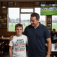 Man and young boy standing together in a sports bar with a TV showing a soccer match.  The boy is wearing a Cows Corner t-shirt with the funny and sweet rugby slogan on the front that reads 'Dad’s My Coach My Biggest Fan and My Post-Match Snack Provider!'. Cows Corner gifts are perfect for sport-mad fans, these gifts work brilliantly for birthdays, new baby celebrations, Father’s Day, Mother’s Day, Christmas, anniversaries, thank you gifts, end-of-season team awards, graduations, retirements
