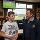 Man and young boy standing together in a pub with a TV showing a sports match in the background. The boy is wearing a Cows Corner t-shirt with the Rugby slogan on the front that reads 'For Every Tackle, Try and Team Huddle – Thanks Coach'. Cows Corner gifts are perfect for sport-mad fans, these gifts work brilliantly for birthdays, new baby celebrations, Father’s Day, Mother’s Day, Christmas, anniversaries, thank you gifts, end-of-season team awards, graduations, retirements, and just-because moments 