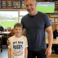 Man and child standing together in a sports bar with rugby-themed decor. The boy is wearing a Cows Corner t-shirt with a funny and sweet rugby slogan on the front that reads 'My Dad Plays Rugby. What’s Your Superpower?'. Cows Corner gifts are perfect for sport-mad fans, these gifts work brilliantly for birthdays, new baby celebrations, Father’s Day, Mother’s Day, Christmas, anniversaries, thank you gifts, end-of-season team awards, graduations, retirements, and just-because moments 