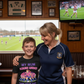 Woman and child in a sports bar watching a rugby match on TV. The boy is wearing a Cows Corner t-shirt with a funny and sweet rugby slogan that reads 'My Mum Scrums Harder Than Your Dad!'. Cows Corner gifts are perfect for sport-mad fans, these gifts work brilliantly for birthdays, new baby celebrations, Father’s Day, Mother’s Day, Christmas, anniversaries, thank you gifts, end-of-season team awards, graduations, retirements, and just-because moments when you want to raise a smile. 
