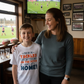Woman and child in a sports-themed room with a TV showing a game. The boy is wearing a Cows Corner t-shirt with the funny and sweet rugby slogan on the front that reads 'My Mum Tackles Everything on the Pitch and at Home!'.  Cows Corner gifts are perfect for sport-mad fans, these gifts work brilliantly for birthdays, new baby celebrations, Father’s Day, Mother’s Day, Christmas, anniversaries, thank you gifts, end-of-season team awards, graduations, retirements, and just-because moments 