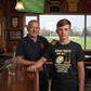 Two men standing in a pub with a television showing a rugby match in the background. The boy is wearing a Cows Corner t-shirt with the Rugby coach slogan on the front that reads 'Rugby Boots Dry Out Coach’s Wisdom Last Forever, Thanks for Everything'. Cows Corner gifts are perfect for sport-mad fans, these gifts work brilliantly for birthdays, new baby celebrations, Father’s Day, Mother’s Day, Christmas, anniversaries, thank you gifts, end-of-season team awards, graduations, retirements, and just-because