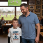 Man and child in a pub watching rugby on TV. The boy is wearing a Cows Corner t-shirt with the funny and sweet Rugby slogan on the front that reads 'Rugby Is Tough but My Dad Is Tougher!'. Cows Corner gifts are perfect for sport-mad fans, these gifts work brilliantly for birthdays, new baby celebrations, Father’s Day, Mother’s Day, Christmas, anniversaries, thank you gifts, end-of-season team awards, graduations, retirements, and just-because moments when you want to raise a smile. 
