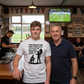 Two men standing together in a pub with a sports bar in the background. The boy is wearing a Cows Corner t-shirt with the rugby coach slogan on the front that reads 'You Taught Me More Than Rugby, You Taught Me Resilience. Thank You, Coach'. Cows Corner gifts are perfect for sport-mad fans, these gifts work brilliantly for birthdays, new baby celebrations, Father’s Day, Mother’s Day, Christmas, anniversaries, thank you gifts, end-of-season team awards, graduations, retirements, and just-because moments