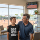 Man and young boy standing together in a room with a tennis court view, wearing a shirt with text.  The boy is wearing a Cows Corner T-shirt with a funny tennis slogan on the front that reads 'My Dads My Coach, My Hero And My Toughest Opponent'.  Cows Corner gifts are perfect for sport-mad fans, these gifts work brilliantly for birthdays, new baby celebrations, Father’s Day, Mother’s Day, Christmas, anniversaries, thank you gifts, end-of-season team awards, graduations, retirements, and just-because moments