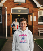 Young boy wearing a hoodie with text in front of a building. The boy is wearing a Cows Corner hoodie with a funny cricket slogan 'Bowled Over by My Mum’s Love & Cooking'. Cows Corner gifts are perfect for sport-mad fans, these gifts work brilliantly for birthdays, new baby celebrations, Father’s Day, Mother’s Day, Christmas, anniversaries, thank you gifts, end-of-season team awards, graduations, retirements, and just-because moments when you want to raise a smile. 
