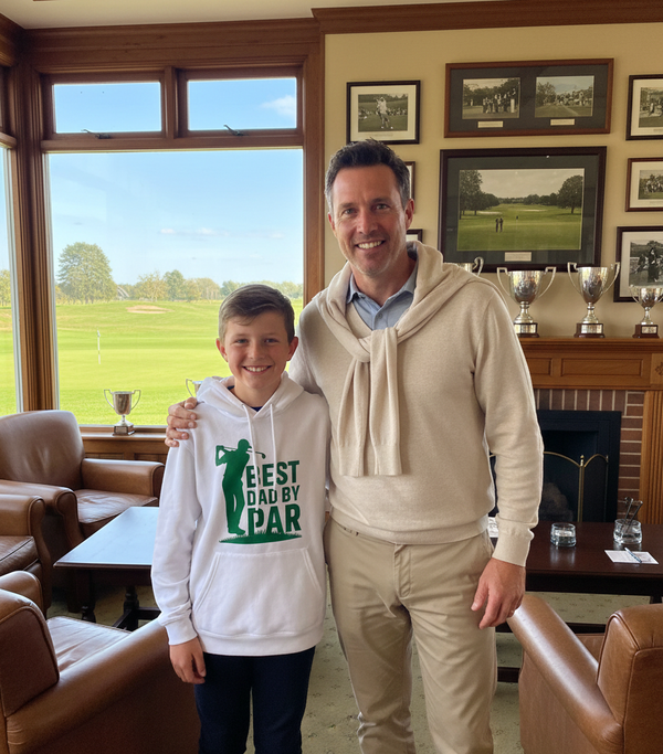 Man and young boy standing together in a room with a view of a golf course. The boy is wearing a Cows Corner hoodie with a funny golf slogan 'Best Dad by Par'. Cows Corner gifts are perfect for sport-mad fans, these gifts work brilliantly for birthdays, new baby celebrations, Father’s Day, Mother’s Day, Christmas, anniversaries, thank you gifts, end-of-season team awards, graduations, retirements, and just-because moments when you want to raise a smile. 
