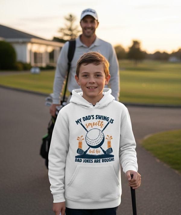 Young boy wearing a hoodie with a golf-themed design, standing on a golf course with a man in the background. The boy is wearing a Cows Corner hoodie with a funny golf slogan 'My Dad’s Swing Is Smooth but His Dad Jokes Are Rough!'. Cows Corner gifts are perfect for sport-mad fans, these gifts work brilliantly for birthdays, new baby celebrations, Father’s Day, Mother’s Day, Christmas, anniversaries, thank you gifts, end-of-season team awards, graduations, retirements, and just-because moments when you want 