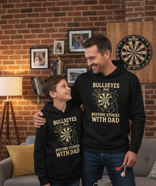 Man and child wearing matching black hoodies with a graphic design and text in a living room setting. The boy is wearing a Cows Corner hoodie with the funny darts slogan on the front that reads 'Bullseyes And Bedtime Stories With Dad'.  Cows Corner gifts are perfect for sport-mad fans, these gifts work brilliantly for birthdays, new baby celebrations, Father’s Day, Mother’s Day, Christmas, anniversaries, thank you gifts, end-of-season team awards, graduations, retirements, and just-because moments 