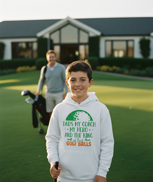 Boy in a hoodie with golf-themed text on a golf course, with a man in the background. A boy wearing a Cows Corner hoodie with a funny golf slogan 'Dads My Coach, My Hero and the King of Lost Golf Balls'. Cows Corner gifts are perfect for sport-mad fans, these gifts work brilliantly for birthdays, new baby celebrations, Father’s Day, Mother’s Day, Christmas, anniversaries, thank you gifts, end-of-season team awards, graduations, retirements, and just-because moments when you want to raise a smile. 
