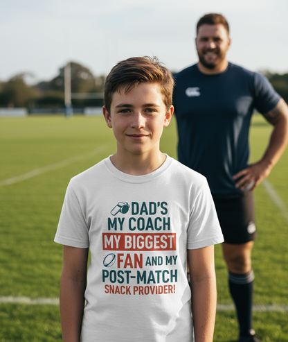 Young boy wearing a t-shirt with a sports-themed message on a soccer field, with an adult in the background. The boy is wearing a Cows Corner t-shirt with the funny rugby slogan 'Dad’s My Coach My Biggest Fan and My Post-Match Snack Provider!'. Cows Corner gifts are perfect for sport-mad fans, these gifts work brilliantly for birthdays, new baby celebrations, Father’s Day, Mother’s Day, Christmas, anniversaries, thank you gifts, end-of-season team awards, graduations, retirements, and just-because moments
