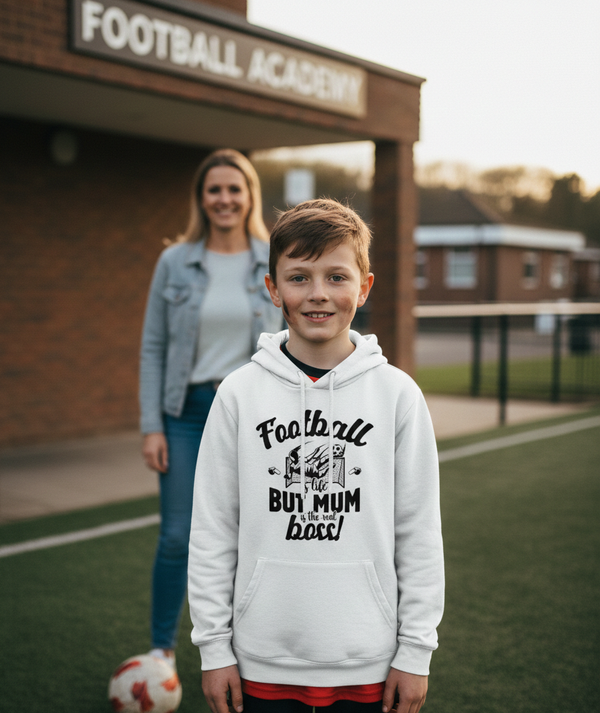 Boy wearing a Cows Corner hoodie with 'Football but mum is the boss' text, standing in front of a football academy. Cows Corner gifts are perfect for sport-mad fans, these gifts work brilliantly for birthdays, new baby celebrations, Father’s Day, Mother’s Day, Christmas, anniversaries, thank you gifts, end-of-season team awards, graduations, retirements, and just-because moments when you want to raise a smile. 
