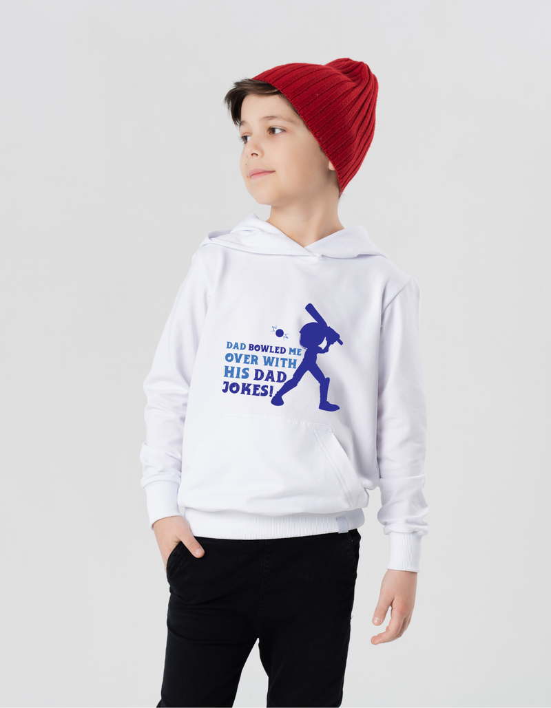 A little boy wearing a white hoodie with Dad Bowled Me Over With His Dad Jokes on the front. A Funny Cricket Hoodie, Premium Fabric, Multiple Junior Sizes, Perfect for Boys and Girls