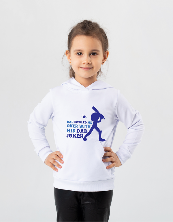 A little girl wearing a white hoodie with Dad Bowled Me Over With His Dad Jokes on the front. A Funny Cricket Hoodie, Premium Fabric, Multiple Junior Sizes, Perfect for Boys and Girls