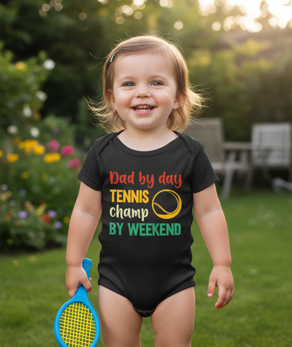 Child wearing a black onesie with a colorful text design, holding a toy tennis racket in a garden setting.  The baby is wearing a Cows Corner baby grow with a funny tennis slogan 'Dad By Day, Tennis Champ By Weekend'. Cows Corner gifts are perfect for sport-mad fans, these gifts work brilliantly for birthdays, new baby celebrations, Father’s Day, Mother’s Day, Christmas, anniversaries, thank you gifts, end-of-season team awards, graduations, retirements, and just-because moments when you want to raise a smi