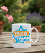 White mug with colorful text and graphics on a wooden table with plants in the background. This is a Cows Corner mug with a funny cricket slogan 'Dad is a Great Fielder, Always Catching Me Out'. Cows Corner gifts are perfect for sport-mad fans, these gifts work brilliantly for birthdays, new baby celebrations, Father’s Day, Mother’s Day, Christmas, anniversaries, thank you gifts, end-of-season team awards, graduations, retirements, and just-because moments when you want to raise a smile. 
