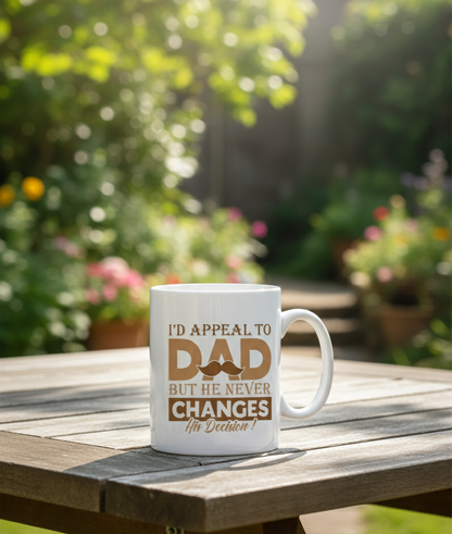 White mug with text on a wooden table outdoors. This is a Cows Corner mug with a funny cricket slogan 'I'd Appeal to Dad, But He Never Changes His Decision'. Cows Corner gifts are perfect for sport-mad fans, these gifts work brilliantly for birthdays, new baby celebrations, Father’s Day, Mother’s Day, Christmas, anniversaries, thank you gifts, end-of-season team awards, graduations, retirements, and just-because moments when you want to raise a smile. 
