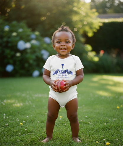Child wearing a 'Dad's Captain' onesie holding a red ball outdoors. The baby is wearing a Cows Corner baby grow with a funny cricket slogan 'Dad's Captain, But I'm The Real Decision-maker'. Cows Corner gifts are perfect for sport-mad fans, these gifts work brilliantly for birthdays, new baby celebrations, Father’s Day, Mother’s Day, Christmas, anniversaries, thank you gifts, end-of-season team awards, graduations, retirements, and just-because moments when you want to raise a smile. 
