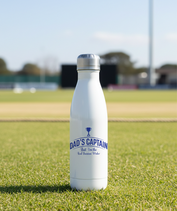 White water bottle with 'Dad's Captain' text on a grassy field. This is a Cows Corner water bottle with a funny cricket slogan 'Dad's Captain, But I'm The Real Decision-maker'. Cows Corner gifts are perfect for sport-mad fans, these gifts work brilliantly for birthdays, new baby celebrations, Father’s Day, Mother’s Day, Christmas, anniversaries, thank you gifts, end-of-season team awards, graduations, retirements, and just-because moments when you want to raise a smile. 
