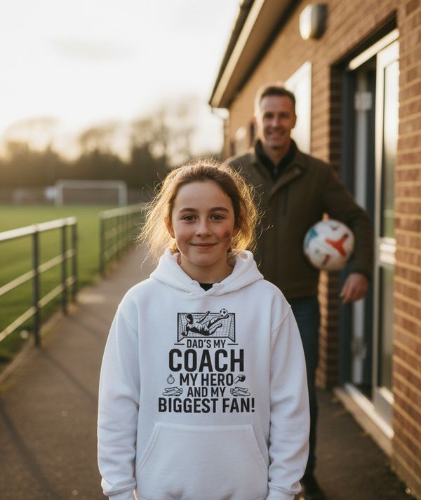 Person wearing a hoodie with text standing in front of a building with another person in the background. The girl is wearing a Cows Corner hoodie with a funny football slogan 'Dad’s My Coach My Hero and My Biggest Fan!'. Cows Corner gifts are perfect for sport-mad fans, these gifts work brilliantly for birthdays, new baby celebrations, Father’s Day, Mother’s Day, Christmas, anniversaries, thank you gifts, end-of-season team awards, graduations, retirements, and just-because moments 