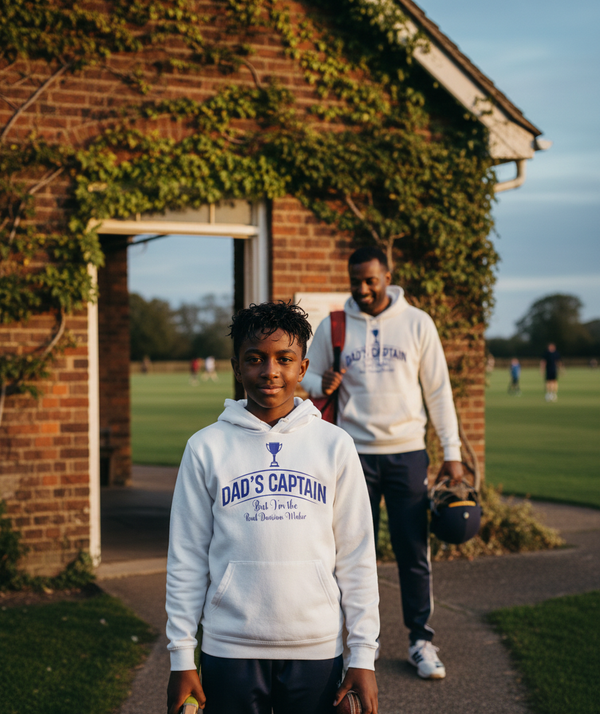 Two individuals wearing 'Dad's Captain' hoodies on a golf course. The boy is wearing a Cows Corner hoodie with a funny cricket slogan 'Dad's Captain, But I'm The Real Decision-maker'. Cows Corner gifts are perfect for sport-mad fans, these gifts work brilliantly for birthdays, new baby celebrations, Father’s Day, Mother’s Day, Christmas, anniversaries, thank you gifts, end-of-season team awards, graduations, retirements, and just-because moments when you want to raise a smile. 
