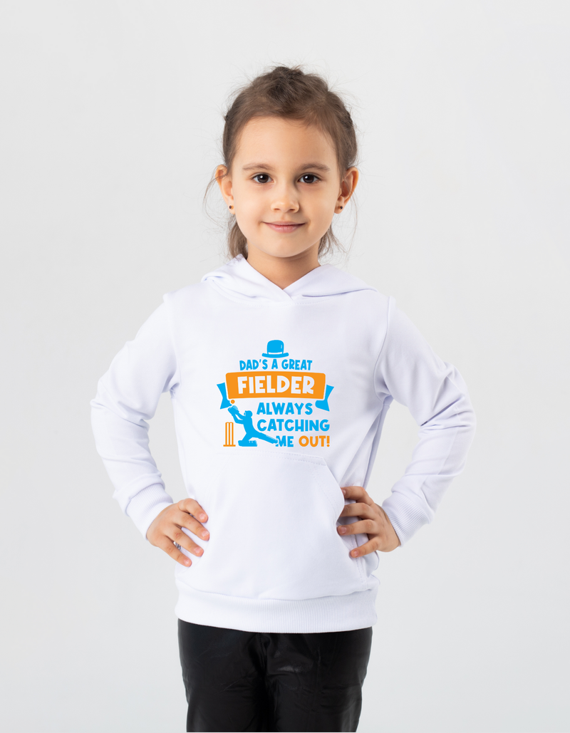 A little girl wearing a white hoodie with Dad Is a Great Fielder, Always Catching Me Out on the front.  A Funny Cricket Hoodie, Premium Fabric, Multiple Junior Sizes, Perfect for Boys and Girls