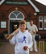 Young boy holding a cricket bat and ball in front of a brick building with 'Willow Creek Cricket Club' sign. The boy is wearing a Cows Corner hoodie with a funny cricket slogan 'Dad Bowled Me Over With His Dad Jokes'. Cows Corner gifts are perfect for sport-mad fans, these gifts work brilliantly for birthdays, new baby celebrations, Father’s Day, Mother’s Day, Christmas, anniversaries, thank you gifts, end-of-season team awards, graduations, retirements, and just-because moments when you want 