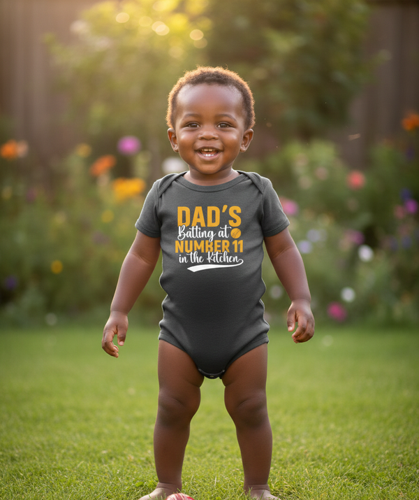 Child wearing a onesie with text in a garden. The baby is wearing a Cows Corner baby grow with a funny cricket slogan 'Dads Batting At Number 11 In The Kitchen'. Cows Corner gifts are perfect for sport-mad fans, these gifts work brilliantly for birthdays, new baby celebrations, Father’s Day, Mother’s Day, Christmas, anniversaries, thank you gifts, end-of-season team awards, graduations, retirements, and just-because moments when you want to raise a smile. 
