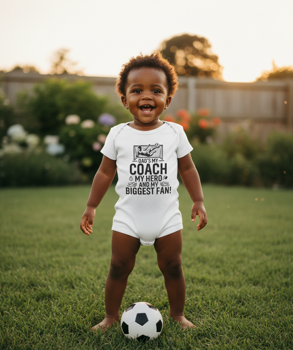Child wearing a white onesie with text on a grassy field. The baby is wearing a Cows Corner baby grow with the funny football slogan 'Dad’s My Coach My Hero and My Biggest Fan!'. Cows Corner gifts are perfect for sport-mad fans, these gifts work brilliantly for birthdays, new baby celebrations, Father’s Day, Mother’s Day, Christmas, anniversaries, thank you gifts, end-of-season team awards, graduations, retirements, and just-because moments when you want to raise a smile. 
