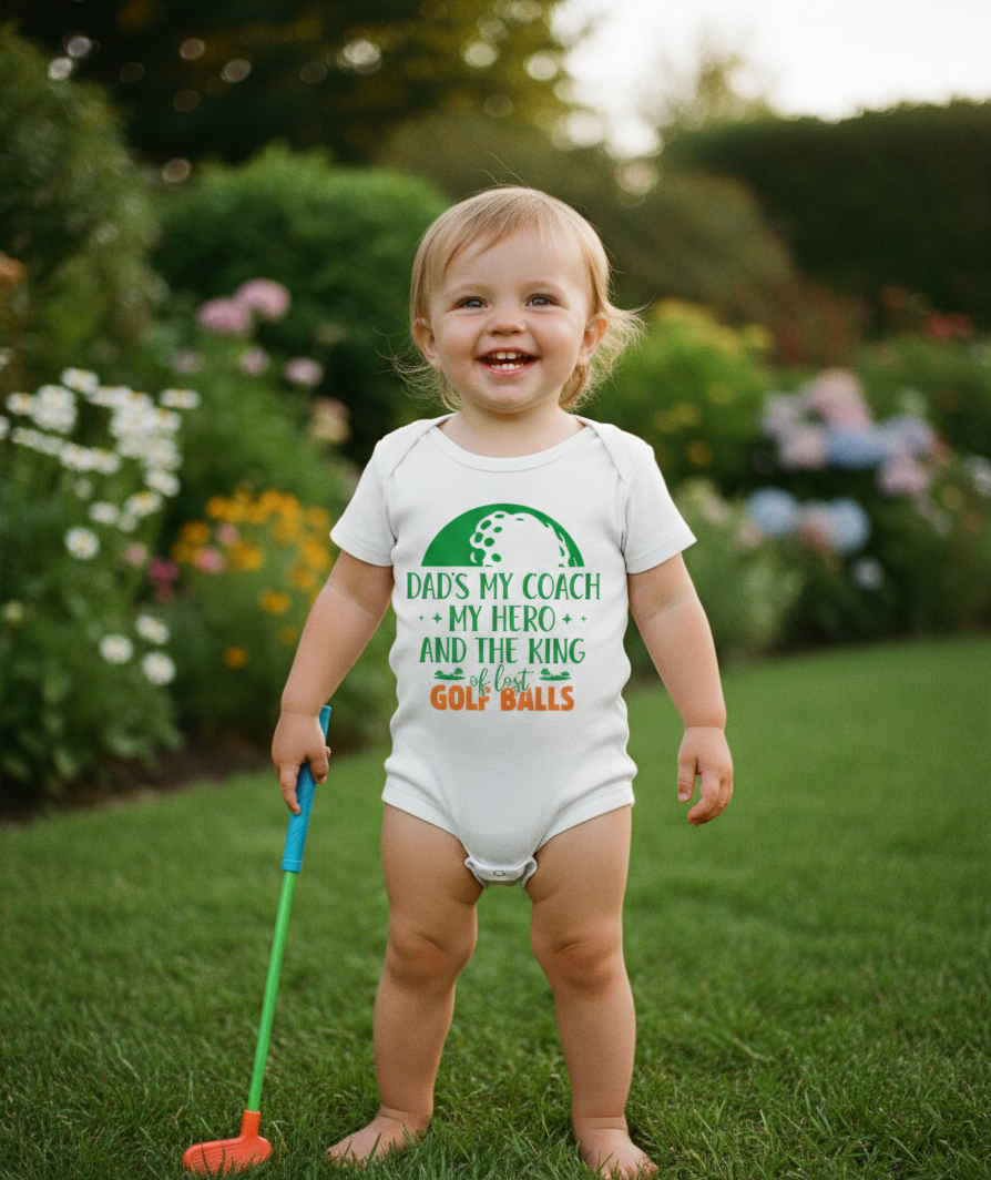 Child wearing a onesie with a golf-themed message, standing outdoors in a garden. The baby is wearing a Cows Corner baby grow with a funny golf slogan 'Dads My Coach, My Hero and the King of Lost Golf Balls'. Cows Corner gifts are perfect for sport-mad fans, these gifts work brilliantly for birthdays, new baby celebrations, Father’s Day, Mother’s Day, Christmas, anniversaries, thank you gifts, end-of-season team awards, graduations, retirements, and just-because moments when you want to raise a smile. 
