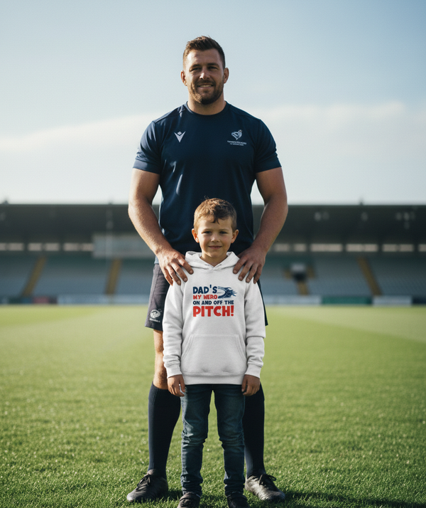 Man and child standing on a sports field with a hoodie text.  The boy is wearing a Cows Corner hoodie with the funny rugby slogan 'Dad’s My Hero on and off the Pitch!'. Cows Corner gifts are perfect for sport-mad fans, these gifts work brilliantly for birthdays, new baby celebrations, Father’s Day, Mother’s Day, Christmas, anniversaries, thank you gifts, end-of-season team awards, graduations, retirements, and just-because moments when you want to raise a smile. 
 