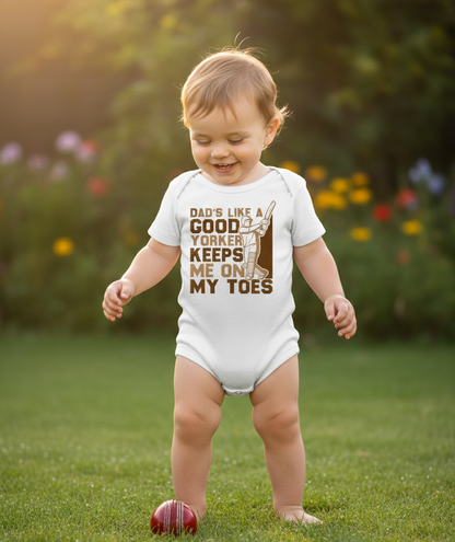 Child wearing a onesie with text in a garden. The baby is wearing a Cows Corner baby grow with a funny cricket slogan 'Dads Like A Good Yorker, Keeps Me On My Toes'. Cows Corner gifts are perfect for sport-mad fans, these gifts work brilliantly for birthdays, new baby celebrations, Father’s Day, Mother’s Day, Christmas, anniversaries, thank you gifts, end-of-season team awards, graduations, retirements, and just-because moments when you want to raise a smile. 

