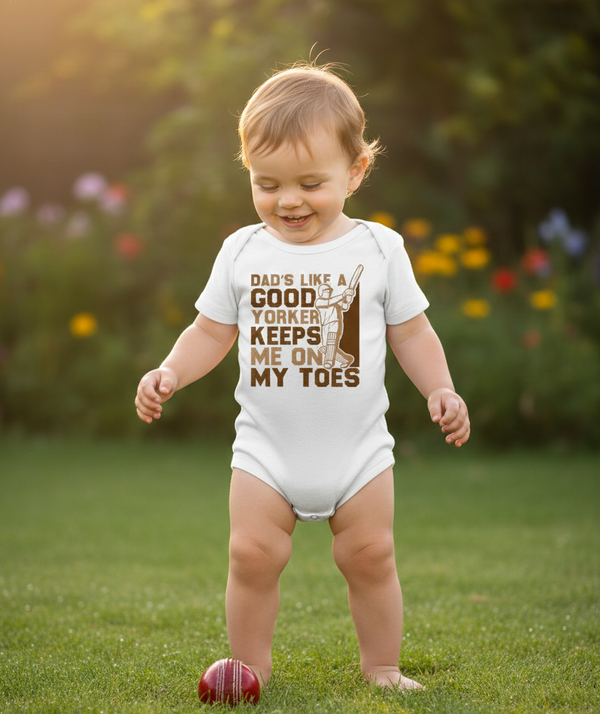 Child wearing a onesie with text in a garden. The baby is wearing a Cows Corner baby grow with a funny cricket slogan 'Dads Like A Good Yorker, Keeps Me On My Toes'. Cows Corner gifts are perfect for sport-mad fans, these gifts work brilliantly for birthdays, new baby celebrations, Father’s Day, Mother’s Day, Christmas, anniversaries, thank you gifts, end-of-season team awards, graduations, retirements, and just-because moments when you want to raise a smile. 
