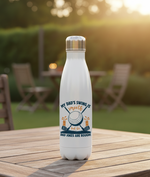 White water bottle with a golf-themed design on a wooden table outdoors. This is a Cows Corner water bottle with a funny golf slogan 'My Dad’s Swing Is Smooth but His Dad Jokes Are Rough!. Cows Corner gifts are perfect for sport-mad fans, these gifts work brilliantly for birthdays, new baby celebrations, Father’s Day, Mother’s Day, Christmas, anniversaries, thank you gifts, end-of-season team awards, graduations, retirements, and just-because moments when you want to raise a smile. 
