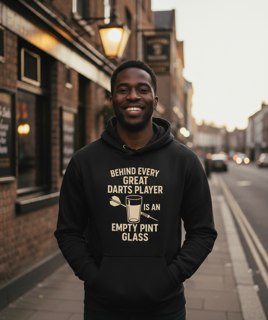 Man wearing a black hoodie with a humorous darts player quote on a street.  The man is wearing a Cows Corner hoodie with a funny darts slogan 'Behind Every Great Darts Player Is An Empty Pint Glass'. Cows Corner gifts are perfect for sport-mad fans, these gifts work brilliantly for birthdays, new baby celebrations, Father’s Day, Mother’s Day, Christmas, anniversaries, thank you gifts, end-of-season team awards, graduations, retirements, and just-because moments when you want to raise a smile. 
