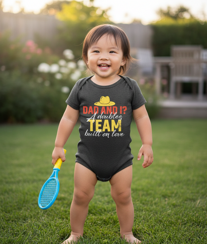 Child wearing a onesie with text outdoors on grass. The baby is wearing a Cows Corner baby grow with a funny tennis slogan 'Dad and I? A Doubles Team Built on Love'. Cows Corner gifts are perfect for sport-mad fans, these gifts work brilliantly for birthdays, new baby celebrations, Father’s Day, Mother’s Day, Christmas, anniversaries, thank you gifts, end-of-season team awards, graduations, retirements, and just-because moments when you want to raise a smile. 
