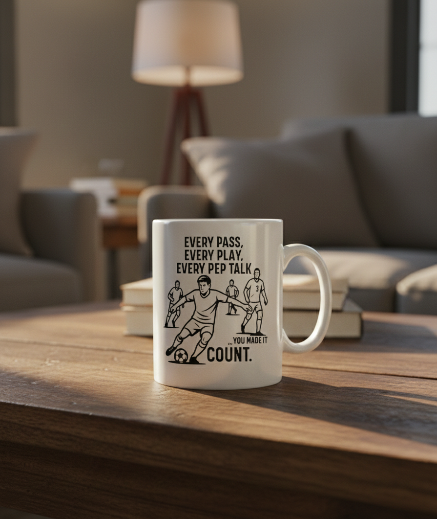 White mug with soccer-themed design and text on a wooden table in a living room setting. This is a Cows Corner mug with a funny football slogan 'Every Pass, Every Play, Every Pep Talk, You Made It Count'. Cows Corner gifts are perfect for sport-mad fans, these gifts work brilliantly for birthdays, new baby celebrations, Father’s Day, Mother’s Day, Christmas, anniversaries, thank you gifts, end-of-season team awards, graduations, retirements, and just-because moments when you want to raise a smile. 
